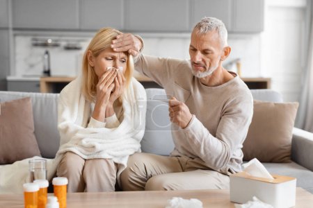 Téléchargez les photos : Homme âgé soignant sa femme malade, tenant un thermomètre et touchant le front de la femme, ayant la grippe saisonnière et le froid, assis sur le canapé à la maison. Fièvre, concept de traitement de la grippe - en image libre de droit