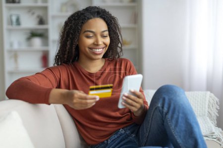 Foto de Happy Young Black Woman Compras en línea con Smartphone y tarjeta de crédito mientras se relaja en el sofá en casa, Mujer afroamericana alegre disfrutando del comercio electrónico, haciendo compras en Internet, Copiar el espacio - Imagen libre de derechos
