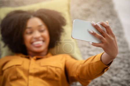 Téléchargez les photos : Femme afro-américaine millénaire souriante aux cheveux bouclés prend un selfie tout en étant couchée, capturant un moment joyeux avec son smartphone dans un environnement confortable, flou - en image libre de droit