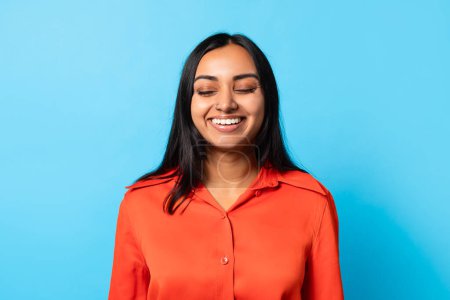 Foto de Felicidad, emociones positivas. Mujer india joven feliz cerrando los ojos y sonriendo, posando para la toma de la cabeza en el fondo del estudio azul, vistiendo camisa naranja, esperando buenas noticias o sorpresa - Imagen libre de derechos