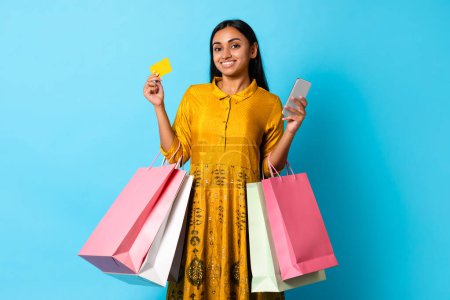 Foto de Un adicto a las compras. Mujer cliente indio feliz sosteniendo Smartphone, tarjeta de crédito y bolsas de compras, haciendo compras a través de la aplicación en el teléfono móvil, mientras que de pie sobre fondo de estudio azul - Imagen libre de derechos