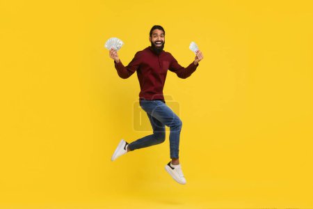 Photo for Cashback, loan, giveaway. Rich wealthy excited handsome millennial indian guy in casual outfit holding money cash dollar banknotes in his hands and jumping, yellow studio background, copy space - Royalty Free Image