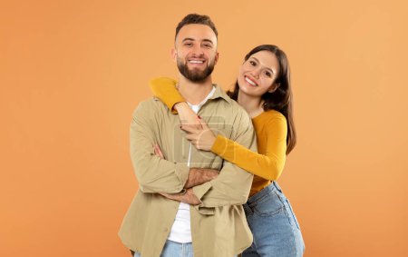 Photo for Romantic Love And Relationship. Smiling European Young Couple Hugging Enjoying Time Together, Standing Over Orange Studio Background, Wife Embracing Her Husband From Back - Royalty Free Image
