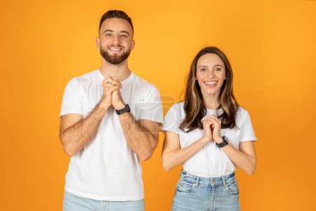Photo for A cheerful european young man and woman in white t-shirts and jeans stand against an orange background, smiling broadly with their hands clasped together in a gesture of excitement and gratitude - Royalty Free Image