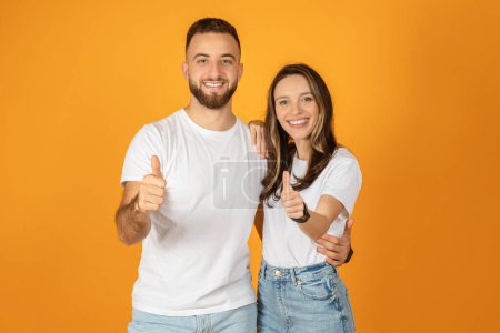 Photo for A beaming happy european young man and woman, dressed in white t-shirts and jeans, enthusiastically give thumbs up to the viewer, against a bright orange background, studio - Royalty Free Image