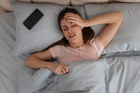 Téléchargez les photos : Jeune femme épuisée éprouvant des maux de tête sévères, pressant son front alors qu'elle était couchée au lit avec un smartphone à côté d'elle, transmettant douleur et inconfort - en image libre de droit