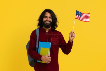 Foto de Estudiante indio sonriente con mochila y un montón de libros que muestran la bandera de los EE.UU. sobre fondo de estudio amarillo, espacio para copiar. Joven positivo oriental que estudia inglés en la escuela. Educación en el extranjero - Imagen libre de derechos