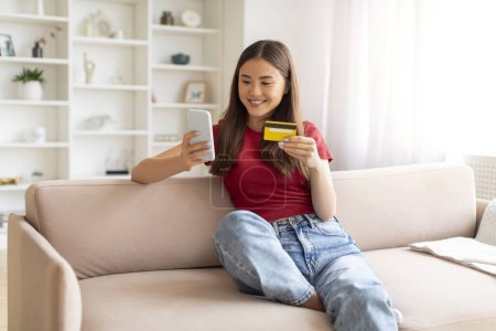Photo for Delighted young asian woman lounging on sofa, engaging in online shopping with her smartphone and credit card, happy korean lady enjoying digital transactions, resting in well-decorated living room - Royalty Free Image