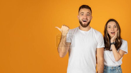 Téléchargez les photos : Un homme européen millénaire choqué dans une chemise blanche pointant avec enthousiasme vers le côté avec une femme surprise réagissant, les deux debout sur un fond orange, panorama - en image libre de droit