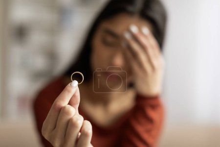 Photo for Upset young woman holding wedding ring in focus with pained expression, depressed millennial female suffering relationship distress or breakup, feeling lonely and stressed, closeup shot - Royalty Free Image