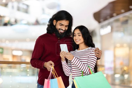 Foto de Comprando juntos Concepto. Retrato de feliz pareja india joven usando smartphone, señora mostrando gadget, chico sosteniendo bolsas de papel de compras, caminando por el centro comercial. Venta al por menor, Venta, Reembolso, Promo - Imagen libre de derechos