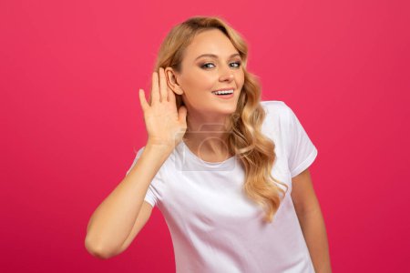 Parle plus fort, concept d'ouïe. Femme blonde joyeuse écoutant tenant la main près de l'oreille sur fond de studio rose, souriant à la caméra. Lady Making Huh Gesture. Je ne t'entends pas.