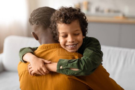 Téléchargez les photos : Joyeux garçon noir aux cheveux bouclés embrasse son père par derrière, son visage montrant un sourire satisfait, exprimant un lien de confiance et d'affection à la maison - en image libre de droit