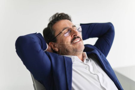 Téléchargez les photos : Relaxé heureux homme d'affaires mature caucasien en costume bleu incliné dans sa chaise avec les mains derrière la tête, les yeux fermés et souriant, prenant une pause dans un espace de bureau lumineux - en image libre de droit
