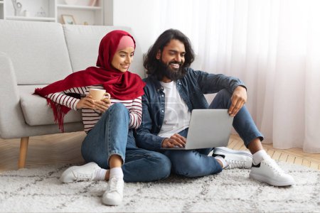 Foto de Relajados pareja joven musulmán sentado en el suelo con ordenador portátil y taza de café, cónyuges islámicos felices utilizando el ordenador juntos mientras se relaja en la acogedora sala de estar, navegar por Internet o ir de compras en línea - Imagen libre de derechos