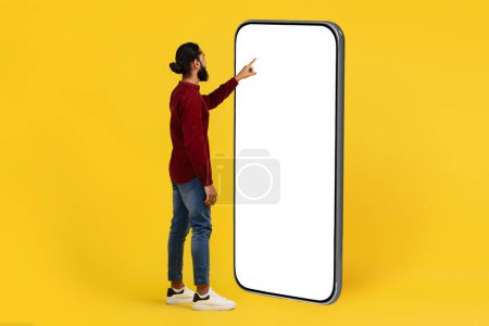 Photo for Cool cheerful bearded young indian man using big phone with white empty touch screen blank copy space mockup for mobile app advertisement, yellow studio background - Royalty Free Image