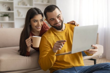 Téléchargez les photos : Surfer sur Internet. Jeune couple se relaxant avec ordinateur portable et café à la maison, joyeux époux millénaire utilisant l'ordinateur et ayant des boissons chaudes dans le salon, profiter week-end domestique, espace libre - en image libre de droit