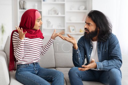 Foto de Conflictos domésticos. Retrato de pareja musulmana joven discutiendo en casa, cónyuges de Oriente Medio que sufren problemas de relación y malentendidos, sentados en el sofá y discutiendo entre sí - Imagen libre de derechos