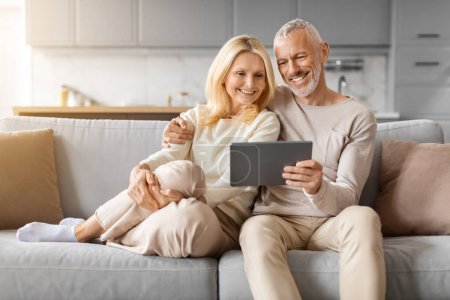 Photo for Positive beautiful loving senior spouses using digital tablet, sitting on couch at home. Happy elderly man and woman paying for goods and services online, using banking or shopping app, copy space - Royalty Free Image
