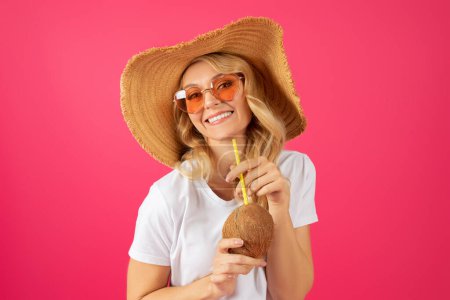 Téléchargez les photos : Portrait d'une jeune femme blonde joyeuse tenant une boisson cocktail à la noix de coco, souriant à la caméra tout en posant dans un chapeau de paille d'été sur fond de studio rose vif, se détendre en vacances ou profiter de la fête - en image libre de droit