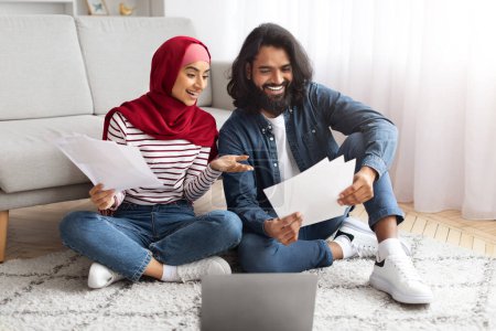 Téléchargez les photos : Joyeux jeunes conjoints musulmans payant des factures sur Internet depuis la maison, assis ensemble sur le sol dans le salon, tenant des papiers financiers, utilisant un ordinateur portable et souriant, profitant facilement e-banking - en image libre de droit