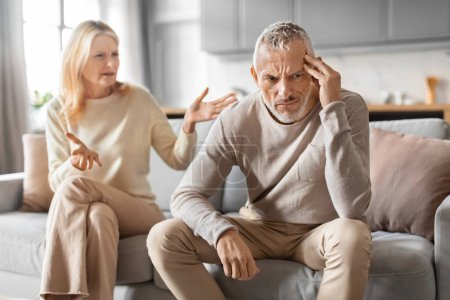 Photo for Furious senior blonde woman yelling at her frustrated irritated husband. Unhappy retied couple sitting on couch at home, have fight, experiencing difficulties in marriage. Crisis in relationships - Royalty Free Image