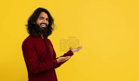 Photo for Cool positive handsome bearded young eastern guy in red shirt pointing at empty copy space and smiling, showing nice offer, deal, recommending nice product, yellow studio background, panorama - Royalty Free Image