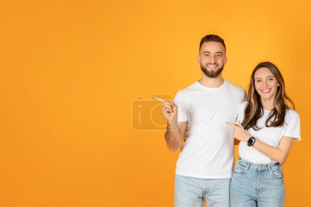 Téléchargez les photos : Un homme et une femme européens joyeux milléniaux en t-shirts blancs et en jeans bleus pointent sur leur côté avec des visages souriants, suggérant un objet ou un concept, sur un fond orange vif - en image libre de droit