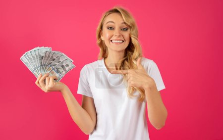 Téléchargez les photos : Jeune femme blonde heureuse tenant de l'argent comptant, pointant du doigt les billets en dollars dans la main sur fond de studio rose, souriant à la caméra, célébrant le succès financier et les bénéfices en espèces - en image libre de droit
