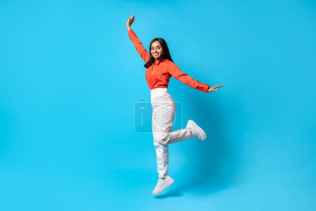 Photo for Joyful carefree Indian young woman jumps and raises arm smiling to camera, posing in mid air over blue studio background. Joy and happiness, celebrating of good news. Empty space, full length - Royalty Free Image