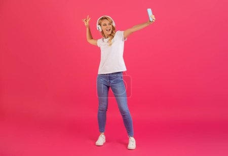 Photo for Cheerful blonde young lady in casual wearing headphones making selfie on smartphone, posing on bright pink studio background, having fun online, full length shot of woman with gadgets, copy space - Royalty Free Image
