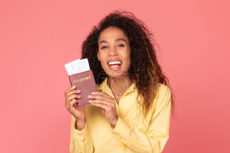 Foto de Mujer negra jubilosa con amplia sonrisa, sosteniendo con confianza su pasaporte y boletos de avión sobre un vibrante fondo rosa, lista para su próxima aventura, disfruta viajar - Imagen libre de derechos