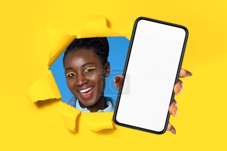 Téléchargez les photos : Offre mobile. Joyeux jeune femme noire montrant son téléphone avec écran vide à travers trou déchiré dans fond de papier jaune, recommandant une nouvelle application ou service. Collage, maquette - en image libre de droit