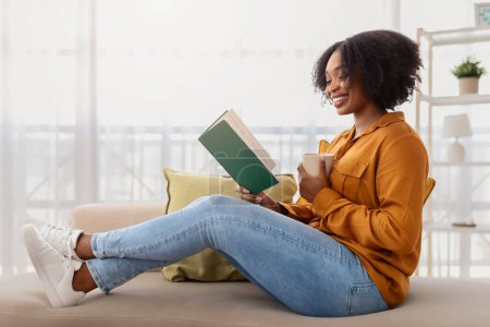 Photo for A relaxed african american woman with curly hair and a radiant smile sits cross-legged on a couch, reading a book and sipping from a cup, in a well-lit, cozy living space - Royalty Free Image
