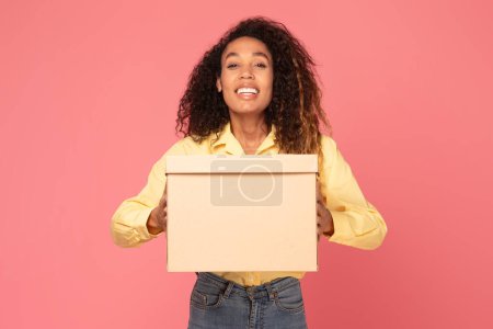 Photo for Cheerful young black woman, exuding happiness, holds box parcel against lively pink background, her smile captures the joy of receiving, making it perfect for conveying positivity and surprise - Royalty Free Image