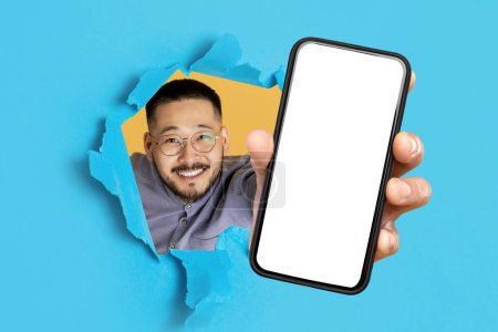 Photo for Smiling japanese businessman showing large smartphone with empty screen through hole in blue paper backdrop, recommending great mobile application, showing mockup for design. Collage - Royalty Free Image