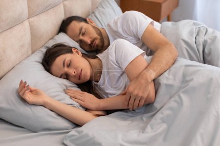 Foto de Pareja en sueño tranquilo, hombre abrazando a la mujer en la cama cómoda con almohadas suaves y sábanas de color azul claro en habitación luminosa - Imagen libre de derechos