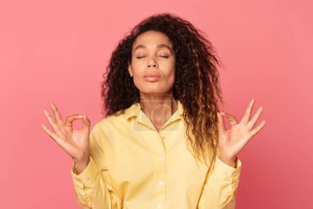 Téléchargez les photos : Femme méditative afro-américaine sereine en chemise jaune avec les yeux fermés et les doigts dans le mudra de yoga, incarnant le calme sur fond rose doux - en image libre de droit