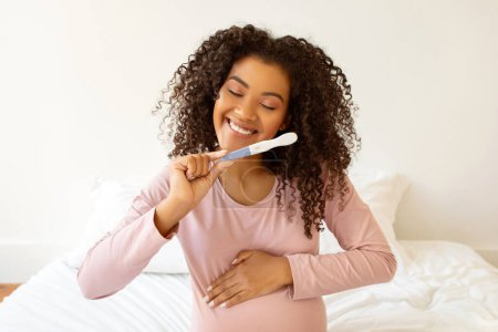 Téléchargez les photos : Rayonnant noir femme enceinte tenant test de grossesse positif et embrassant son ventre, heureuse jeune femme enceinte afro-américaine anticipant le voyage à venir, profiter de la maternité à venir - en image libre de droit