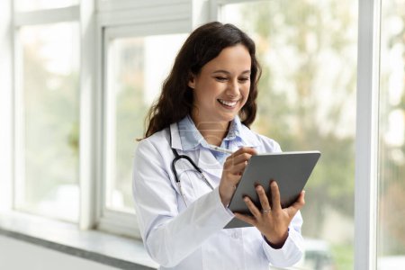 Téléchargez les photos : Une jeune femme souriante portant un manteau médical médecin debout à côté de la fenêtre à la clinique, à l'aide d'une tablette numérique, a rendez-vous en ligne avec le patient. Télésanté, assurance-maladie à distance - en image libre de droit