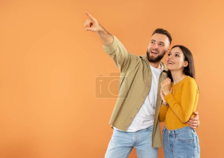 Photo for Nice Offer. Excited young caucasian man pointing finger aside at empty ad place hugging woman, showing something to his wife, offering empty space for text on orange studio background - Royalty Free Image