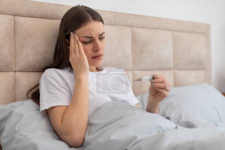 Photo for Concerned young woman lying in bed, holding her head and looking at thermometer, her face showing worry over possible fever or illness, copy space - Royalty Free Image
