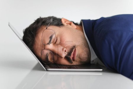 Photo for An overworked caucasian businessman in a blue suit takes a nap on his laptop keyboard in an office setting, a clear sign of fatigue, burnout, or working long hours, close up - Royalty Free Image
