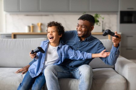 Foto de Muchacho preadolescente negro exuberante y su padre compartiendo momento alegre, riendo y jugando videojuegos juntos, creando recuerdos en su acogedora sala de estar - Imagen libre de derechos
