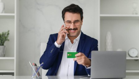 Téléchargez les photos : Homme d'affaires mature caucasien souriant en costume bleu parlant au téléphone et tenant une carte de crédit verte, effectuant éventuellement une transaction, avec un ordinateur portable sur son bureau, panorama - en image libre de droit