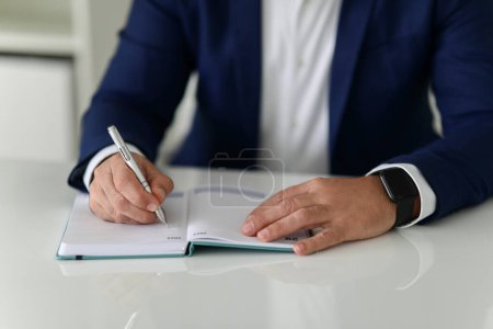 Téléchargez les photos : Gros plan d'un homme d'affaires caucasien écrivant dans un carnet, portant une montre intelligente et un costume bleu, sur une table blanche dans un bureau bien éclairé, recadré. Travail, entreprise - en image libre de droit