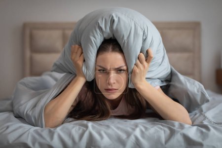 Téléchargez les photos : Jeune femme agacée en pyjama utilisant un oreiller pour bloquer le bruit, les yeux fermés, montrant un regard de frustration alors qu'elle était allongée dans son lit - en image libre de droit