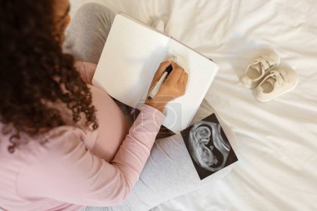 Téléchargez les photos : La future mère écrit dans un journal à côté d'une échographie de bébé et de petites chaussures, une jeune femme noire méconnaissable remplissant son journal de grossesse, capturant de précieux moments d'anticipation, ci-dessus - en image libre de droit
