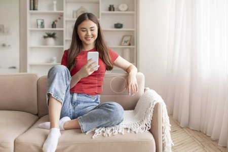 Téléchargez les photos : Relaxé jeune femme asiatique assis sur le canapé et regardant son smartphone, souriant coréen femelle avec téléphone portable dans les mains se prélasser sur le canapé dans le salon, messagerie avec des amis ou la navigation nouvelle application - en image libre de droit