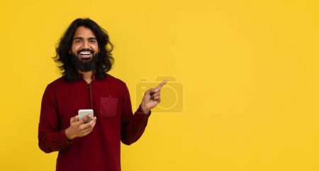 Téléchargez les photos : Positif joyeux élégant jeune homme indien utilisant un smartphone, pointant vers l'espace de copie vierge, recommandant une belle application mobile, bannière web, isolé sur fond de studio jaune - en image libre de droit